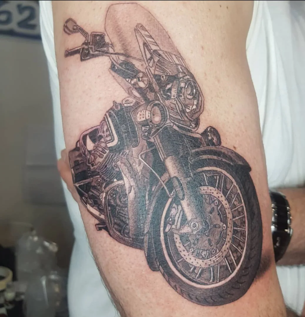 Tatouage réaliste d'une moto en noir et gris sur le bras, détails de mécanique et chrome, réalisé par Jean-Marie Joullié au salon JMJ Tattoo à Béziers et Sérignan